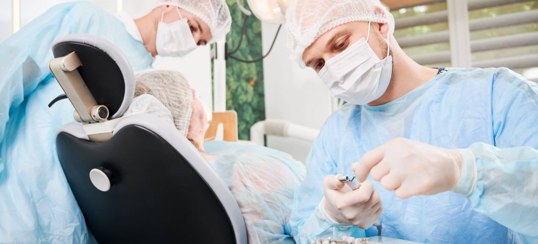 Dentist on foreground, wearing disposable sterile clothes, putting surgical nozzle on drill before implant installation. Patient and assistant on blurred background. Concept of implant placement.