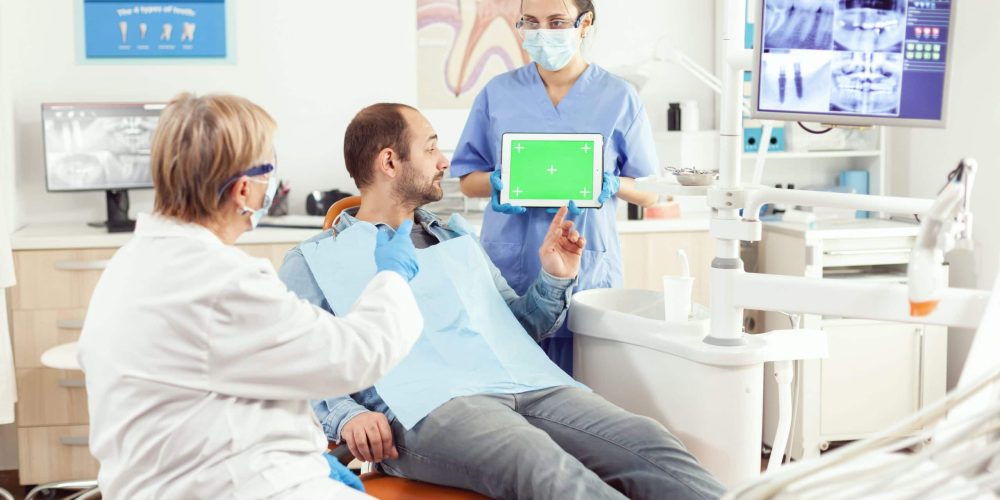 Medical nurse holding mock up green screen chroma key tablet with isolated display during somatology consultation. Sick patient sitting on dental chair waiting for somatology x-ray scan