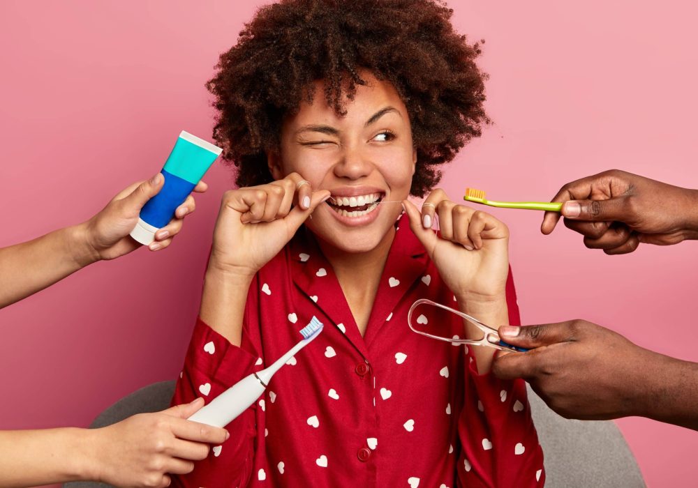 Oral hygiene and teeth care concept. Curly haired woman cleans teeth with floss, cleans tongue with cleaner, uses toothbrush and toothpaste, poses at home, wears nightwear, has perfect healthy tooth
