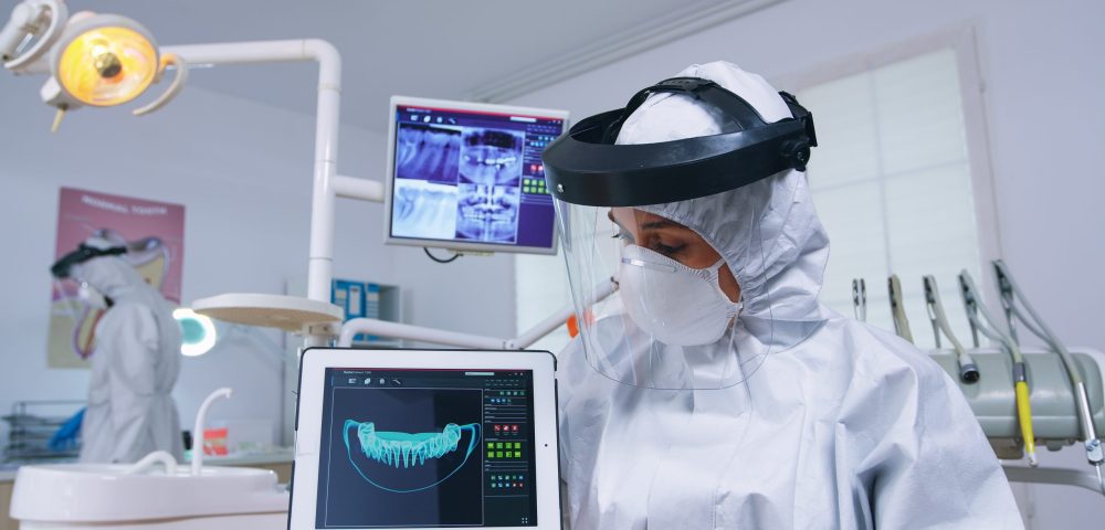 Patient pov listening explication for teeth treatment, dentist in coverall showing x-ray on tablet. Stomatology specialist wearing protective suit against infection with covid19 pointing at radiography