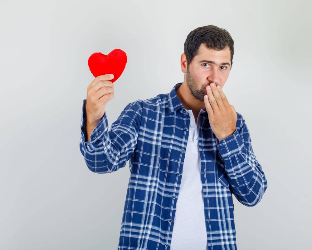 Young man holding red heart with hand on mouth in shirt and looking shy , front view.